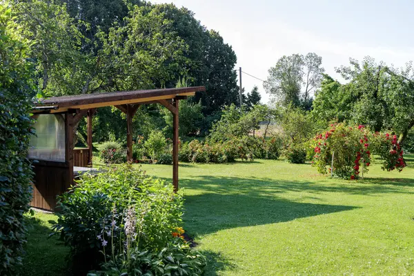 Canlı yeşillikler, güneşli çimenler ve uzakta çiçek açan güllerle çevrili ahşap bir teras ya da kulübe içeren huzurlu bir bahçe sahnesi..