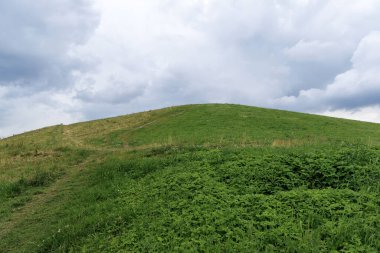 Çerçeveye yemyeşil bir tepe hükmediyor, sola doğru kıvrılan bir yol gösteriyor, hepsi uçsuz bucaksız, parçalı bulutlu gökyüzünün altında.