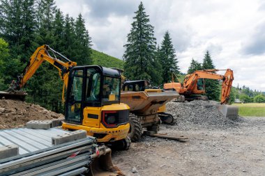  Orman ve çakıllarla çevrili bir dağ manzarasında, bir inşaat sahasına park edilmiş mini bir kazıcı ve çöplük..