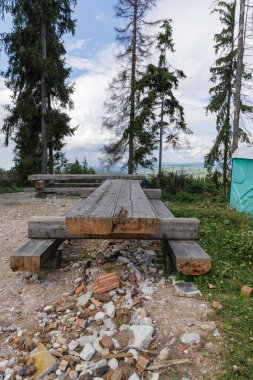 Basit bir ahşap piknik masası ve dağlık bir arazide, bulutlu bir gökyüzünün altındaki çam ağaçlarıyla çevrili banklar. Yapı enkazı görünür..