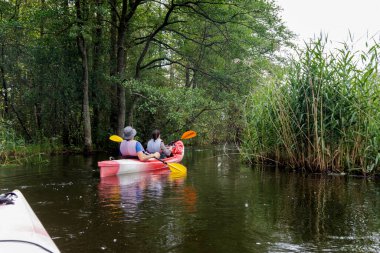 Bir tarafında sık bir orman, diğer tarafında uzun kamışlı iki kişinin kürek çektiği bir kayık görüntüsü..