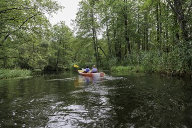 İki kişi, sakin, yeşil bir nehirde, sık ormanlı ve kıyılarda sazlıklar olan bir kanoyla kameradan uzaklaşır..