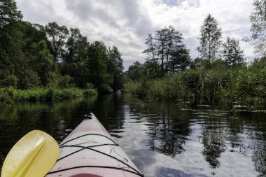 Dolambaçlı, yeşil bir nehirde yüzen bir kanonun önünden başka bir kano bulutlu bir gökyüzünün altında görünür..
