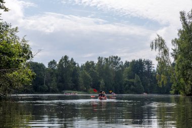 Bir grup kanocu, bulutlu bir gökyüzünün altında yemyeşil bir ormanın oluşturduğu geniş ve sakin bir nehirde kürek çekiyor..