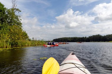  Bir grup kanocu, yeşil kıyıları olan geniş ve sakin bir nehirde, dağılmış bulutlarla dramatik bir gökyüzünün altında, özgürlük ve beraberlik hissini yakalıyorlar..