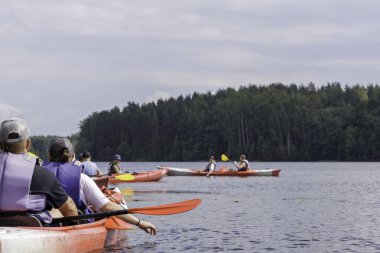Bir kanonun arkasından yakın çekim görüntüsü, bulutlu bir gökyüzünün altındaki geniş bir nehirde iki kişinin diğer kayakçılarla kürek çektiğini gösteriyor..