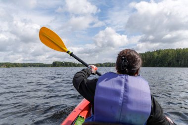 Bir insan, geniş ve dalgalı bir gölde, açık mavi gökyüzünün altında, tüylü beyaz bulutları olan bir orman kıyısına doğru kano sürüyor..