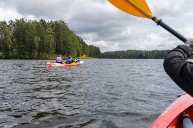 Bir kanonun manzarası, bulutlu bir gökyüzünün altındaki ormanlık bir göle doğru birlikte kürek çeken iki arkadaşı takip ediyor..