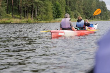 Bir kanonun manzarası, iki insanın birlikte kürek çektiğini gösteriyor. Uzak kıyılarda yemyeşil bir orman var..