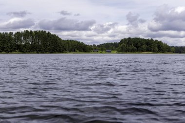 Dalgalanan bir gölün geniş ve geniş bir manzarası, uzak bir orman kıyısı ve dramatik bulutlu bir gökyüzünün altında iki küçük bina..