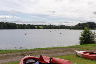 Çimenli sahilde iki boş kano dinlenirken, bir grup kanocu uzaktaki sakin suda kürek çekiyor..