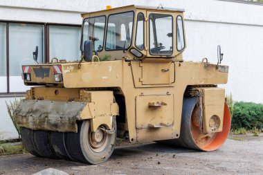  Geniş trampetli eski, sarı ve siyah bir yol silindiri beyaz bir binaya karşı bir caddeye park edilmiş..