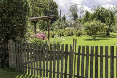 Kırsal bir ahşap çitin içindeki açık bir kapı çiçek açan bitkiler, çimenler ve meyve ağaçları olan yeşil bir arka bahçeye çıkar..
