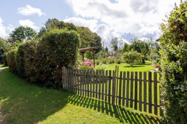 Eski ahşap bir çit yeşil bir çimenlikte gölgeler bırakır, çalılar ve çiçekli bir bahçeye çıkar..