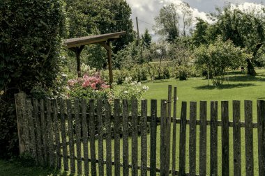 Yıpranmış ahşap bir çitin ve kapının yakın çekimi kırsal dokusunu gözler önüne seriyor ve huzurlu, yeşil bir bahçeye götürüyor..