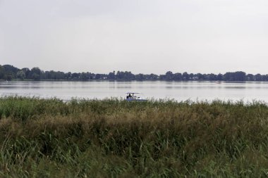 Yolcularla dolu küçük motorlu bir tekne orta mesafede geniş ve sakin bir gölde, ön planda uzun kamışların arkasında görülür..