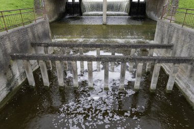  Ön planda köpük ve türbülans yaratan büyük dikey filtre kolonları ve şelale sızıntısını gösteren çok katlı beton baraj yapısı.