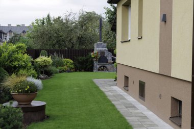 Yeşil bir çimenlik, asfalt kaldırım, manikürlü çalılar ve arka planda kırsal taştan bir fırın bulunan modern bir banliyö evinin temiz ve bakımlı bahçesi.. 