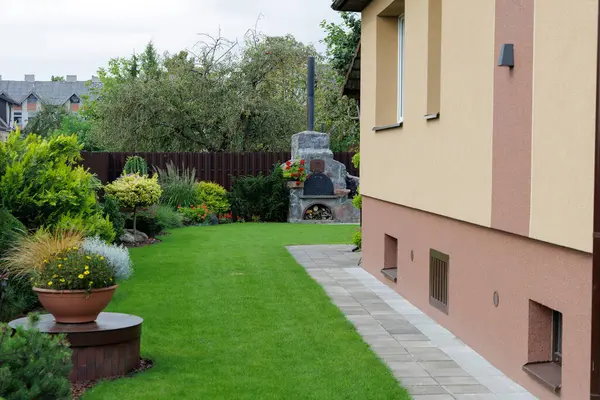 Yeşil bir çimenlik, asfalt kaldırım, manikürlü çalılar ve arka planda kırsal taştan bir fırın bulunan modern bir banliyö evinin temiz ve bakımlı bahçesi.. 