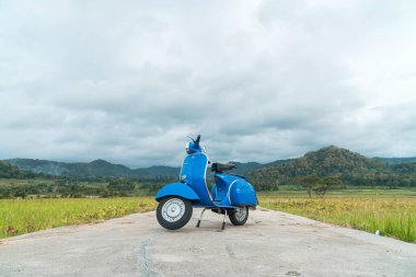 Temiz ve parlak mavi klasik bir Vespa yolun ortasına park etmiş yeşil pirinç tarlasında arka planda tepeler var.