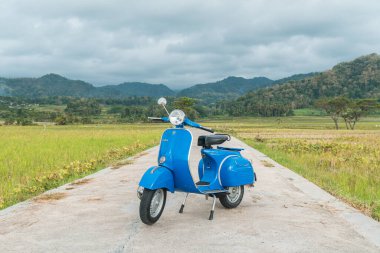 Temiz ve parlak mavi klasik bir Vespa yolun ortasına park etmiş yeşil pirinç tarlasında arka planda tepeler var.