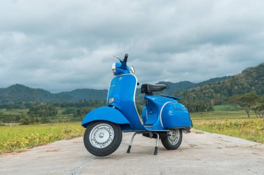 Temiz ve parlak mavi klasik bir Vespa yolun ortasına park etmiş yeşil pirinç tarlasında arka planda tepeler var.