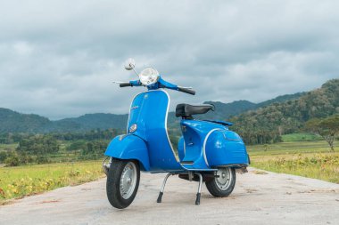 Temiz ve parlak mavi klasik bir Vespa yolun ortasına park etmiş yeşil pirinç tarlasında arka planda tepeler var.