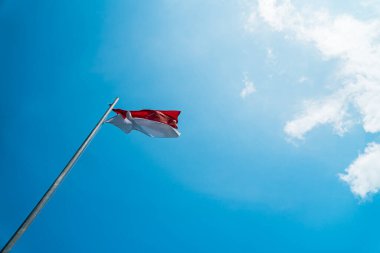 Red and white flag, Indonesia's national flag flying above the pole for independence day in a bright blue sky with white clouds around it
