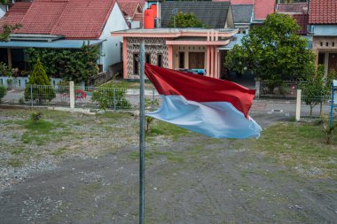 Red and white flag, Indonesia's national flag flying above the pole for independence day in a bright blue sky with white clouds around it