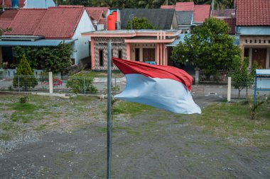 Red and white flag, Indonesia's national flag flying above the pole for independence day in a bright blue sky with white clouds around it