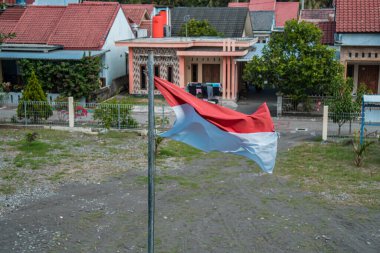 Red and white flag, Indonesia's national flag flying above the pole for independence day in a bright blue sky with white clouds around it