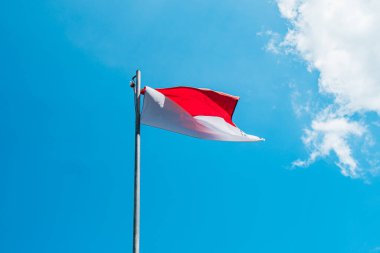 Red and white flag, Indonesia's national flag flying above the pole for independence day in a bright blue sky with white clouds around it