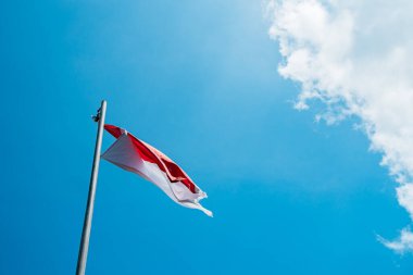Red and white flag, Indonesia's national flag flying above the pole for independence day in a bright blue sky with white clouds around it