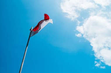 Red and white flag, Indonesia's national flag flying above the pole for independence day in a bright blue sky with white clouds around it