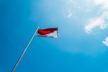 Red and white flag, Indonesia's national flag flying above the pole for independence day in a bright blue sky with white clouds around it