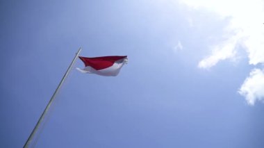 Red and white flag, Indonesia's national flag flying above the pole for independence day in a bright blue sky with white clouds around it