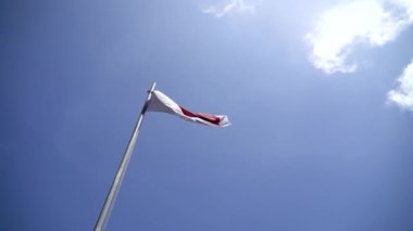 Red and white flag, Indonesia's national flag flying above the pole for independence day in a bright blue sky with white clouds around it