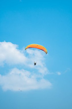 Adrenalin salgılayan bir ekstrem spor, açık mavi gökyüzüne karşı yamaç paraşütü. Gökyüzünde paraşütüyle uçan paraşütçü. Arka planda mavi gökyüzü ve güneşli bir günde beyaz bulutlar..