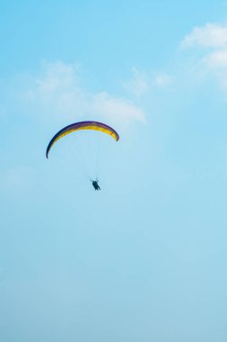 Adrenalin salgılayan bir ekstrem spor, açık mavi gökyüzüne karşı yamaç paraşütü. Gökyüzünde paraşütüyle uçan paraşütçü. Arka planda mavi gökyüzü ve güneşli bir günde beyaz bulutlar..