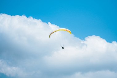 Adrenalin salgılayan bir ekstrem spor, açık mavi gökyüzüne karşı yamaç paraşütü. Gökyüzünde paraşütüyle uçan paraşütçü. Arka planda mavi gökyüzü ve güneşli bir günde beyaz bulutlar..