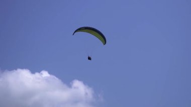 Adrenalin salgılayan bir ekstrem spor, açık mavi gökyüzüne karşı yamaç paraşütü. Gökyüzünde paraşütüyle uçan paraşütçü. Arka planda mavi gökyüzü ve güneşli bir günde beyaz bulutlar..