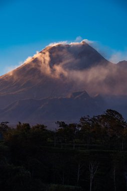 Karanlık çökmeden Merapi Dağı 'nın güzelliği ve önünde soğuk lav uçurumu var. Merapi Dağı açık bir günde mavi gökyüzü ve yanında bulutlarla detaylı görünüyor.