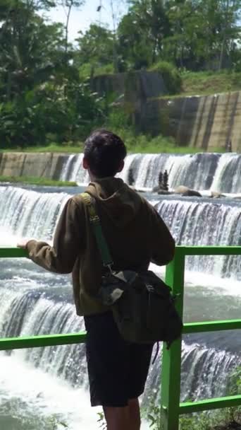 サボダムの人工滝で一人で休暇をするアジア人男性は 6つのレベルを持っています 男の後ろの景色は 水の6つのレベルを持っているサボダムの美しいパノラマを楽しんでいます — ストック動画