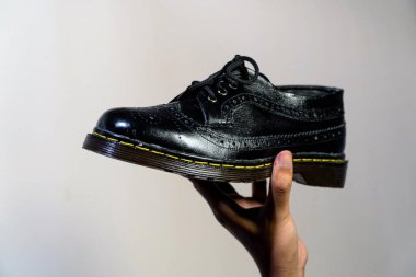 A man's hand holds a full black brogue wingtip shoe with a rubber outsole made from genuine cowhide. Men's hands holding elegant and shiny vintage shoe on a cream background clipart