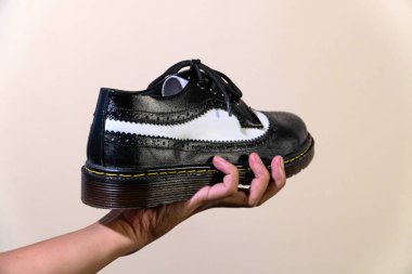 Bir adamın eli, hakiki sığır derisinden yapılmış kauçuk tabanlı siyah beyaz kanat uçlu bir ayakkabı tutar. Erkek eli, krem rengi arka planda zarif ve parlak iki renkli ayakkabılar tutuyor.