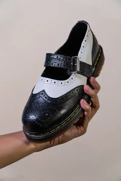 Bir adamın eli, hakiki sığır derisinden yapılmış kauçuk tabanlı siyah beyaz Mary Jane Rockabilly ayakkabısını tutuyor. Erkek eli, krem rengi arka planda zarif ve parlak iki renkli ayakkabılar tutuyor.