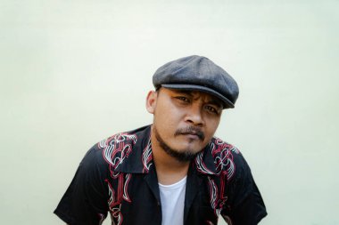 Sakallı ve bıyıklı maço adam bej arka planda çok yalnız görünüyor. Eski moda bir Güneydoğu Asyalı yetişkin adamın yarı vücut portresi desenli gömlek, beyaz atlet bluz ve gazeteci çocuk şapkası.