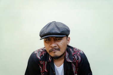 Sakallı ve bıyıklı maço adam bej arka planda çok yalnız görünüyor. Eski moda bir Güneydoğu Asyalı yetişkin adamın yarı vücut portresi desenli gömlek, beyaz atlet bluz ve gazeteci çocuk şapkası.