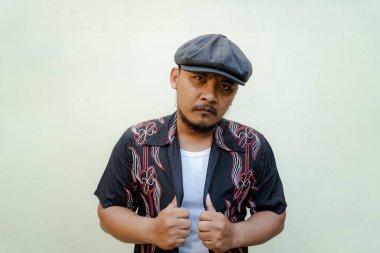 Sakallı ve bıyıklı maço adam bej arka planda çok yalnız görünüyor. Eski moda bir Güneydoğu Asyalı yetişkin adamın yarı vücut portresi desenli gömlek, beyaz atlet bluz ve gazeteci çocuk şapkası.