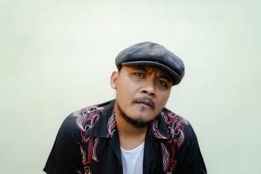 Sakallı ve bıyıklı maço adam bej arka planda çok yalnız görünüyor. Eski moda bir Güneydoğu Asyalı yetişkin adamın yarı vücut portresi desenli gömlek, beyaz atlet bluz ve gazeteci çocuk şapkası.